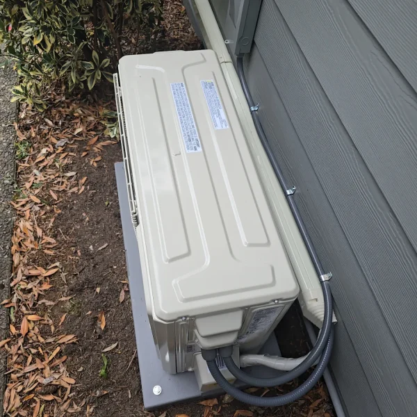 Mini-split outdoor unit on a mounting pad beside grey siding, with disconnect box and line set
